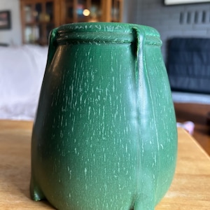 May include: A forest green ceramic vessel with a rounded, bulbous body and a textured, weathered finish. The container features a thick, raised rim at the top and three small, integrated feet at the base, providing a stable, elevated stance.