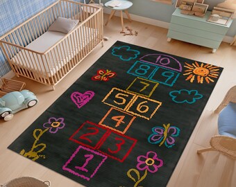 May include: A black rectangular rug with a colourful hopscotch design. The rug features a hopscotch grid with numbers 1-10, along with drawings of a heart, flowers, butterflies, a sun, and clouds. The rug is placed on a light wood floor.