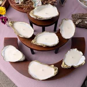 May include: Handmade oyster shell candles with gold leaf trim. The candles are white and sit in real oyster shells. The shells are arranged on a wooden display stand. Other shell-shaped items are in the background.
