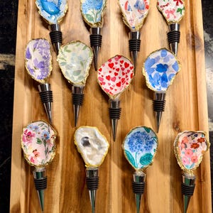 May include: A collection of decorative wine bottle stoppers. Each stopper features a unique design on an oyster shell, including floral patterns, hearts, and nautical themes. The shells have gold-colored edges and are set on black and silver stoppers.