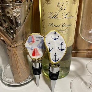 May include: Two decorative wine bottle stoppers. One stopper features a nautical design with blue sailboats and red sails. The other stopper has a white background with blue anchors. Both stoppers have a gold trim and a silver base.