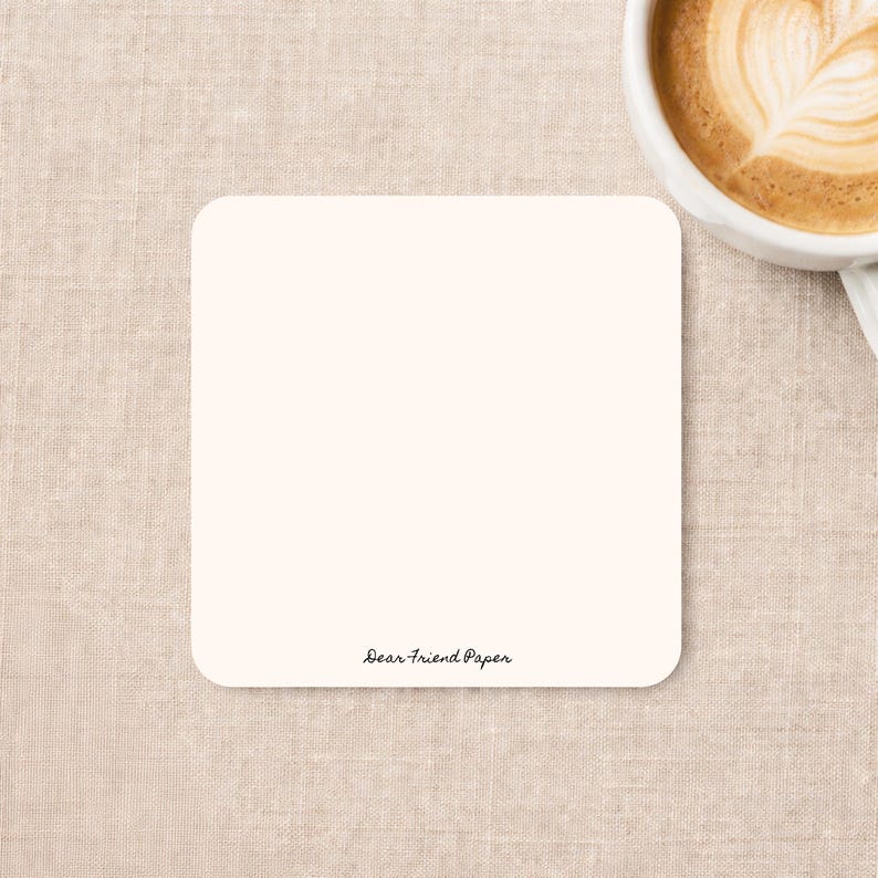May include: A square, off-white coaster with rounded corners. The text "Dear Friend Paper" is printed in a small, black, cursive font near the bottom center of the coaster. The surface of the coaster is smooth and solid in color.