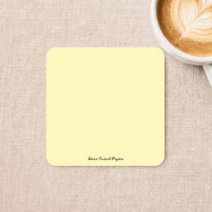 May include: A square, pale yellow notepad with rounded corners. The bottom center of the notepad features the text Dear Friend Paper in a small, black, cursive font. The notepad is positioned next to a white ceramic mug filled with coffee.