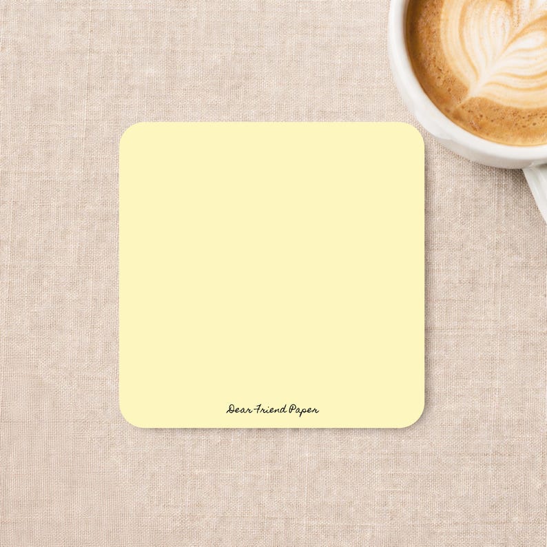 May include: A square, pale yellow notepad with rounded corners. The bottom center of the notepad features the text Dear Friend Paper in a small, black, cursive font. The notepad is positioned next to a white ceramic mug filled with coffee.