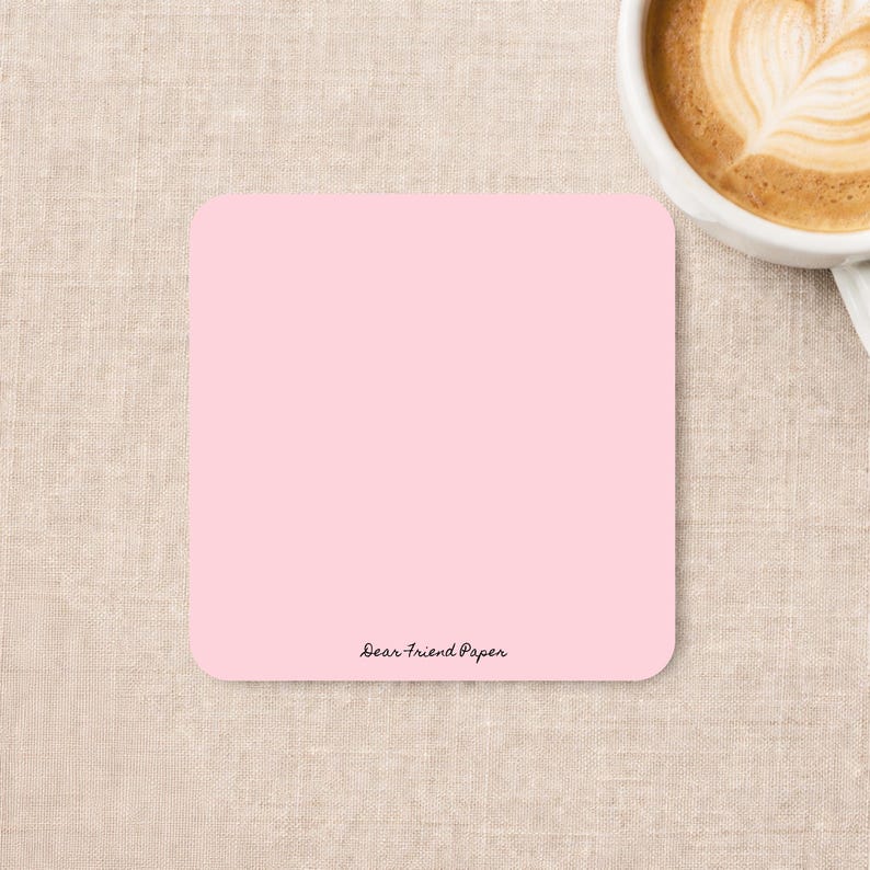 May include: A square, solid light pink coaster with rounded corners. The text "Dear Friend Paper" is printed in a small, black, cursive font near the bottom center of the coaster. The surface of the coaster is smooth and matte in appearance.