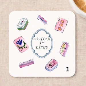 May include: A square coaster featuring a watercolor illustration of various Mahjong tiles in pink, green, and blue hues surrounding a central scalloped frame. The text inside the frame reads Mahjong at Kate's. The number 1 is printed in black.