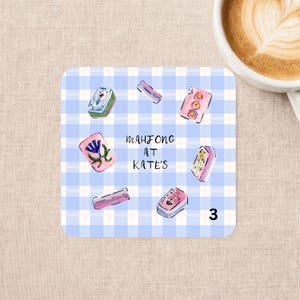 May include: A square coaster with a light blue and white gingham pattern featuring illustrations of various mahjong tiles arranged in a circle. The text "MAHJONG AT KATE'S" is printed in the center in a simple black handwritten font.