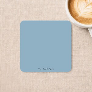 May include: A square coaster in a solid dusty blue color with rounded corners. The text "Dear Friend Paper" is printed in a small, black, cursive font near the bottom center of the coaster surface. The coaster has a smooth, matte finish.