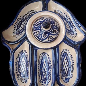 Peut inclure: Un plat de service en céramique blanche en forme de main avec cinq compartiments en forme de doigts. Le plat est décoré de motifs géométriques bleus et blancs et possède un couvercle avec un design similaire.