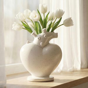 May include: A heart-shaped, speckled ceramic vase filled with white tulips. The vase has a unique design with a textured surface and a decorative top. The flowers have green stems and leaves, creating a fresh and elegant floral arrangement.