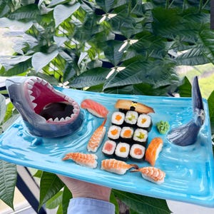 May include: A blue ceramic serving tray shaped like a shark with a dipping bowl in the shark's mouth. The tray has a variety of sushi rolls, nigiri, and sashimi arranged on it. The tray also has a small ceramic shark fin on the right side.