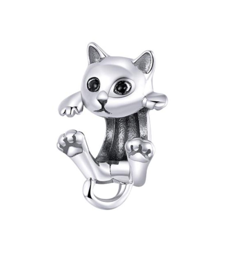 May include: A silver-colored cat-shaped jewelry piece. The cat has large, dark eyes and is posed as if it is hanging or climbing. The design is detailed, with visible paws and a curled tail, creating a playful and whimsical aesthetic.