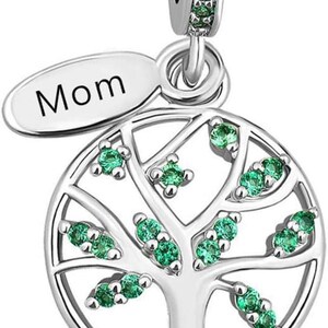 Könnte beinhalten: Ein silberner Charm mit einem kreisförmigen Lebensbaum-Design und grünen Steinen in den Ästen. Ein ovaler Anhänger an der oberen Schlaufe zeigt das Wort Mom in schwarzer Schrift. Die Öse ist ebenfalls mit kleinen grünen Steinen besetzt.
