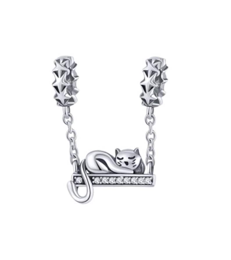 May include: A silver-colored charm featuring a sleeping cat on a swing. The swing is adorned with small, clear stones and hangs from two star-shaped connectors. The cat is curled up, with its tail gently curved.
