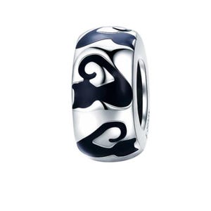 May include: A silver-colored charm with a cylindrical shape. The charm features a repeating pattern of black, swirling designs. The design is set against a shiny, metallic background. The charm is likely for a bracelet or necklace.