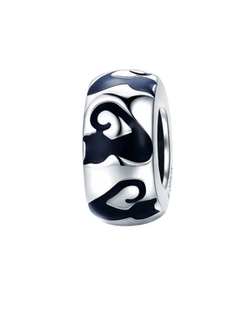 May include: A silver-colored charm with a cylindrical shape. The charm features a repeating pattern of black, swirling designs. The design is set against a shiny, metallic background. The charm is likely for a bracelet or necklace.