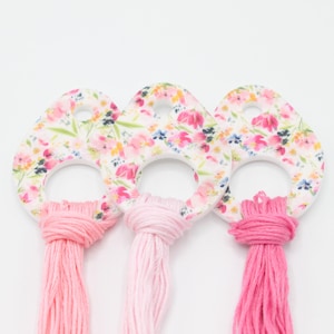 May include: Three floral-patterned, teardrop-shaped wooden keychains with pink tassels. The keychains have a white background with a pattern of pink, red, and blue flowers. The tassels are different shades of pink.