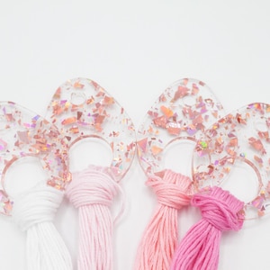 May include: Clear, teardrop-shaped resin pendants with embedded pink and iridescent glitter, paired with white, light pink, pink, and hot pink embroidery floss. These pendants are ideal for crafting and jewelry making.