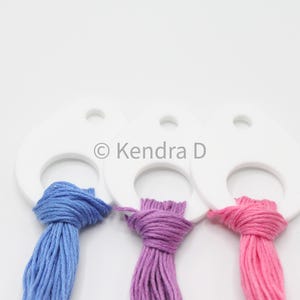May include: Three white, teardrop-shaped thread organizers with blue, purple, and pink embroidery floss. Each organizer has a hole at the top and a larger opening at the bottom to hold the floss. The text "© Kendra D" is printed on the organizers.