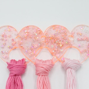 May include: A heart-shaped craft organizer in clear resin, filled with pink glitter and small embellishments. Three bundles of pink embroidery floss are attached. The organizer is designed to hold and display thread.