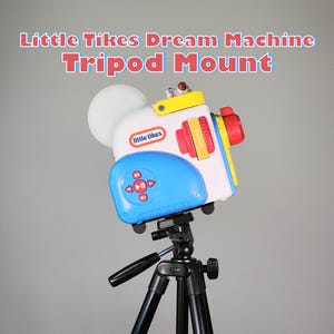 May include: A colorful Little Tikes Dream Machine Tripod Mount toy. The toy is white, blue, yellow, and red, with a large white lens and a control panel. It is mounted on a black tripod.