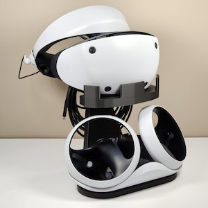 May include: A white VR headset with a black stand and two controllers. The headset is on top of the stand, and the controllers are on the bottom.