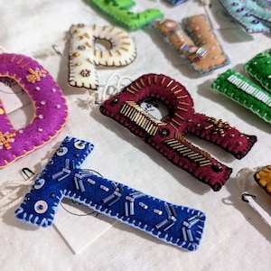 May include: A set of felt letter ornaments, each decorated with colorful beads and sequins. The letters are in various colors, including green, white, brown, purple, red, blue, and light blue. The letters are all uppercase and are arranged in a random order.