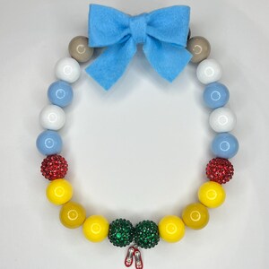May include: A colorful beaded necklace with a blue felt bow, red and green sparkly beads, and a charm of red shoes. The necklace is made of large, round beads in shades of white, blue, yellow, and red.