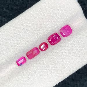 May include: Five faceted loose gemstones in varying shades of vibrant pink and red. The collection includes one small rectangular stone, one larger rectangular stone, one round stone, and two cushion-cut stones, all arranged in a single line.