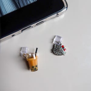 Könnte beinhalten: Zwei transparente Handy-Anhänger aus Kunststoff mit goldenen Ringen. Ein Anhänger zeigt eine Comicfigur mit einer roten Schleife und einem silbernen Glitzerdesign. Der andere Anhänger zeigt einen braunen und weißen Comic-Boba-Tee mit einem schwarzen Strohhalm.