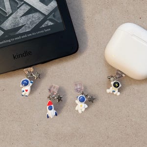 May include: A collection of astronaut-themed charms with star accents, attached to a Kindle and a white AirPods case. The charms feature miniature astronauts and a rocket ship in white, blue, and red. The Kindle is black, and the AirPods case is white.