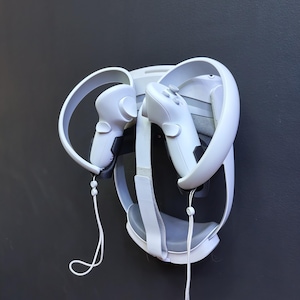 May include: A white VR headset and controllers are mounted on a dark grey wall. The headset has a curved design with a strap and the controllers have buttons and hand straps. The setup is designed for virtual reality gaming.