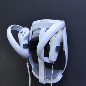 Puede incluir: Auriculares y mandos de realidad virtual blancos montados en una pared gris oscuro. Los auriculares tienen un cojín gris y correas blancas. Los mandos tienen botones y bucles para correas de muñeca. El conjunto está diseñado para juegos de realidad virtual.