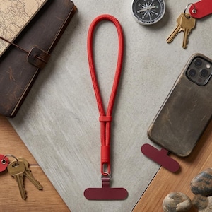 May include: A red phone lanyard with a matching phone grip is displayed on a neutral surface. The lanyard is made of a woven material and has a loop for the wrist. Other items include a leather journal, keys, a compass, and a phone case.