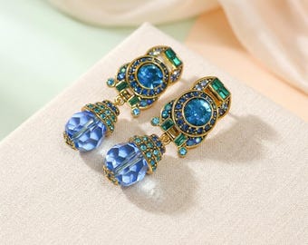 May include: A pair of ornate earrings featuring a gold-tone setting with blue and green gemstones. Each earring has a round, blue center stone surrounded by smaller blue and green gems. A faceted blue teardrop hangs below.