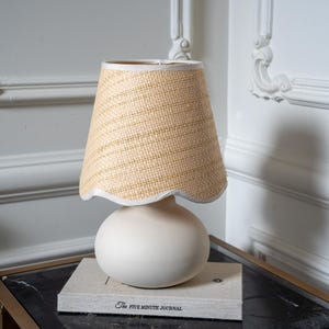 May include: A table lamp with a smooth, matte white spherical base and a textured, light tan woven lampshade. The lampshade features a scalloped bottom edge with a white trim. The lamp rests on a book titled The Five Minute Journal.