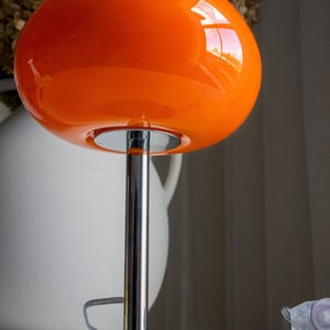 May include: A vibrant orange table lamp with a rounded shade and a chrome-finished stem and base. The lamp's glossy shade reflects light, and the design is reminiscent of mid-century modern aesthetics.