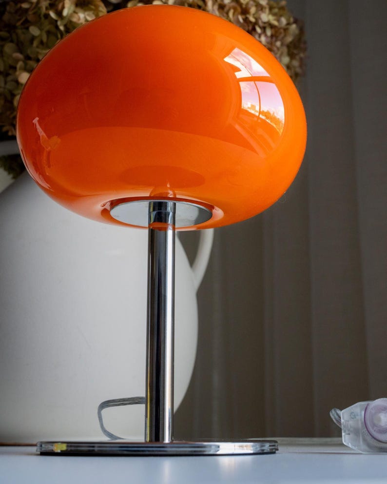 May include: A vibrant orange table lamp with a rounded shade and a chrome-finished stem and base. The lamp's glossy shade reflects light, and the design is reminiscent of mid-century modern aesthetics.