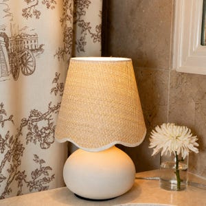 May include: A table lamp with a smooth, matte white rounded base and a textured, light beige woven lampshade. The lampshade features a scalloped bottom edge with a white trim, providing a soft, diffused light source for any interior space.