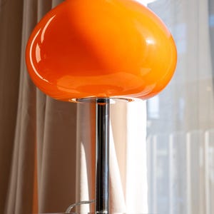 May include: A retro-style table lamp with a vibrant orange, mushroom-shaped glass shade. The lamp features a chrome-finished, cylindrical stem and a circular base. The design is reminiscent of mid-century modern decor.
