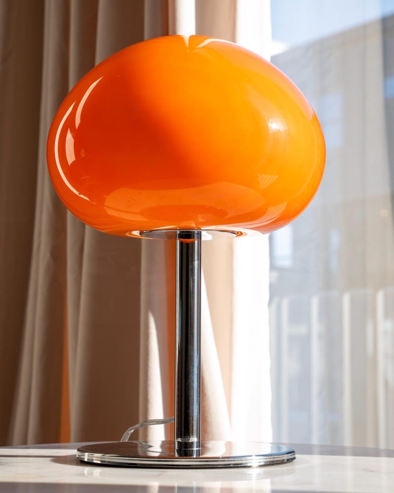 May include: A retro-style table lamp with a vibrant orange, mushroom-shaped glass shade. The lamp features a chrome-finished, cylindrical stem and a circular base. The design is reminiscent of mid-century modern decor.