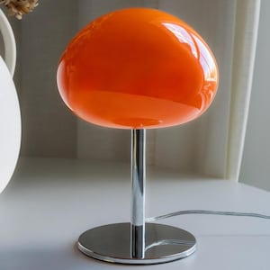 May include: A retro-style table lamp with a vibrant orange glass shade. The shade is round and sits atop a chrome-finished stem and circular base. The lamp has a shiny, reflective surface and a power cord.
