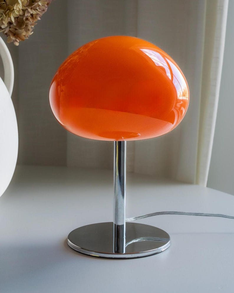 May include: A retro-style table lamp with a vibrant orange glass shade. The shade is round and sits atop a chrome-finished stem and circular base. The lamp has a shiny, reflective surface and a power cord.