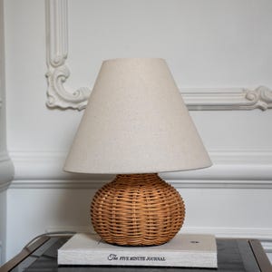 May include: A table lamp with a round, woven rattan base and a conical, off-white fabric lampshade. The lamp sits on top of a rectangular book titled The Five Minute Journal, which features a light gray cover with black text.