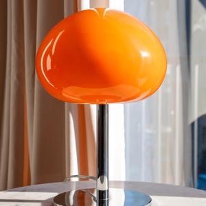 May include: An orange mushroom-shaped table lamp with a chrome base and stem. The lamp has a glossy, rounded shade and a circular base. The lamp is sitting on a white marble table. The background includes a sheer white curtain.