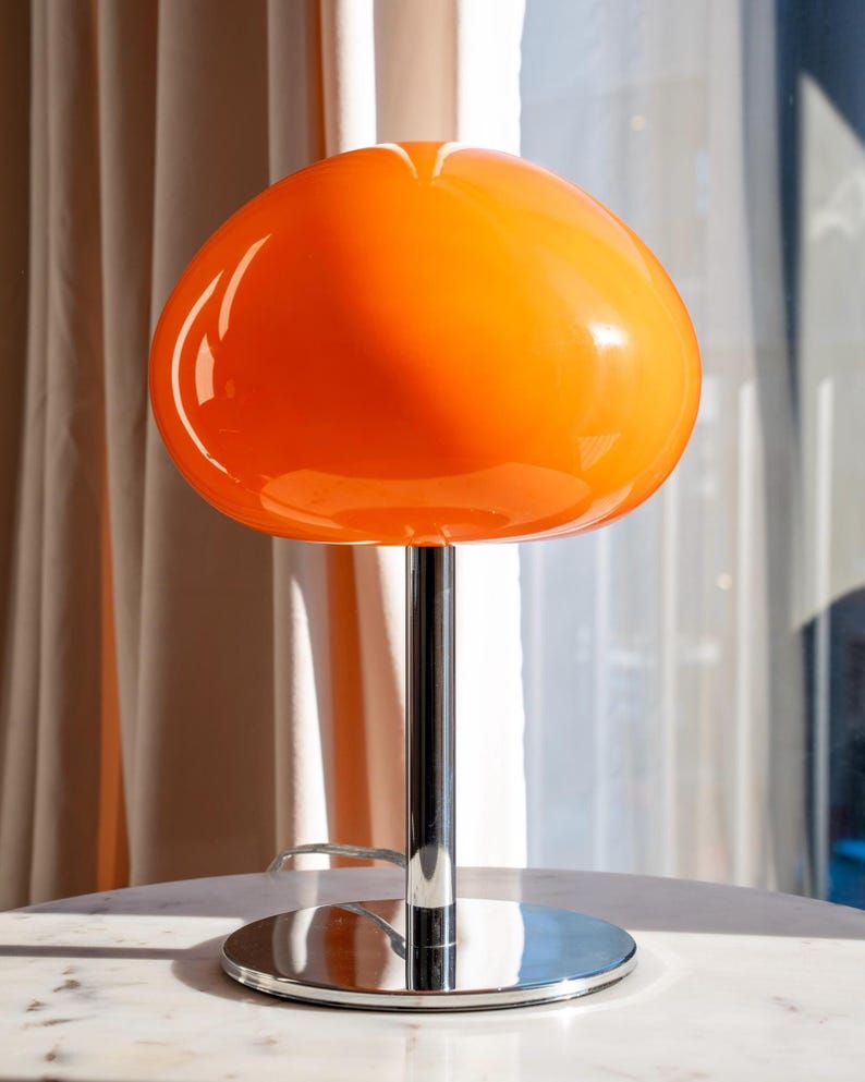 May include: An orange mushroom-shaped table lamp with a chrome base and stem. The lamp has a glossy, rounded shade and a circular base. The lamp is sitting on a white marble table. The background includes a sheer white curtain.
