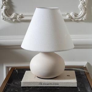 May include: A table lamp with a matte cream-colored spherical base and a white conical fabric lampshade. The lamp rests on a rectangular beige book titled The Five Minute Journal, which features black text on its front cover.