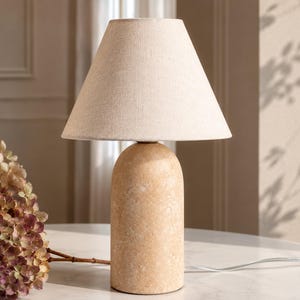 May include: A beige table lamp with a textured, cylindrical base and a cream-colored lampshade. The lamp has a simple, modern design and a neutral color palette. The lamp is on a white surface.