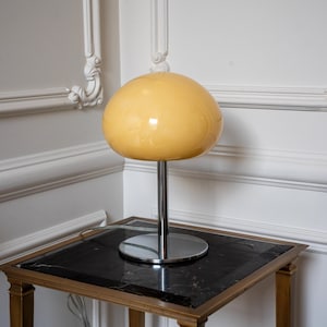 May include: A table lamp featuring a smooth, glossy, pale yellow mushroom-shaped glass shade. The lamp is supported by a slender, polished chrome metal stem that connects to a circular, flat, reflective chrome base, creating a sleek profile.
