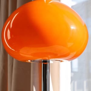May include: A vibrant orange, mid-century modern style lamp with a large, rounded shade. The shade sits atop a chrome-colored, slender pole. The lamp's design is reminiscent of a mushroom shape, casting a warm glow.